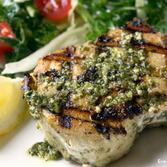 A piece of grilled halibut with pesto sauce, garnished with a lemon and served with salad.