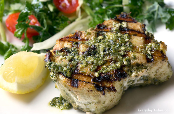A piece of grilled halibut with pesto sauce, garnished with a lemon and served with salad.