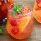 Two glasses of a peach white wine sangria, with fresh peaches and raspberries, and garnished with mint.