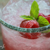 A glass full of a non-alcoholic raspberry spritzer that's garnished with fresh raspberries and mint.