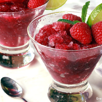 Two glasses of a mango raspberry margarita granita, garnished with fresh raspberries and lime.