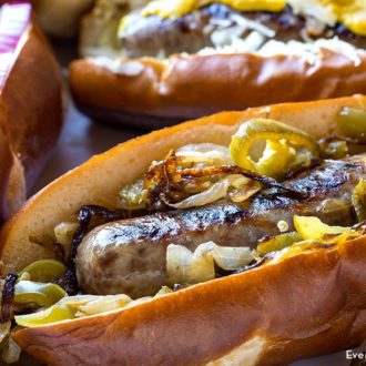 A grilled beer brat in a bun and garnished with veggies.
