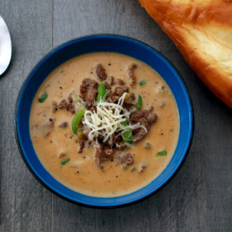 A bowl of hearty Philly cheesesteak stew, topped with shredded cheese, with a loaf of bread — a perfect dinner on a cold night