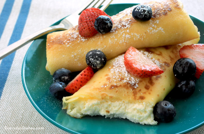 Two homemade cheese blintzes on a plate and topped with powdered sugar and fresh fruit.