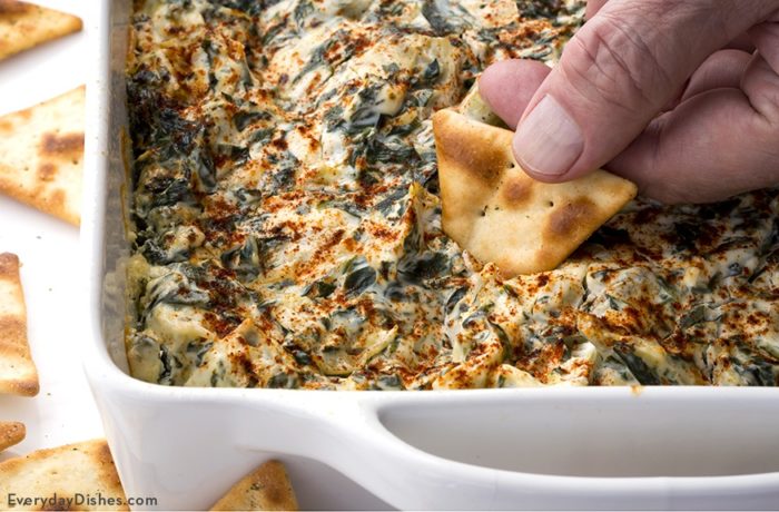 A dish of homemade simple spinach artichoke dip with a hand dipping a cracker into it.